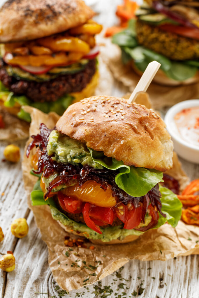 Vegan burger, carrot burger, homemade burger with carrot cutlet, grilled bell pepper, cherry tomatoes, red onion chutney, lettuce, and avocado sauce,on a white, wooden table. Healthy eating concept, healthy alternative